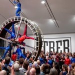 Dancers suspend in a giant wheel with large audience surrounding them