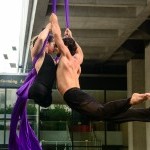 color photo of a muscular male and female aerialist spinning from purple silks, the female aerialist on the left has no legs