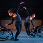 Two dancers wearing black and grey bodysuits, standing beside two wheelchairs side by side on a blue lighted floor.