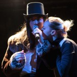 Two performers lean their faces towards a microphone, one in a top hat and fur coat, the other in a black dress.