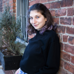 Sophie, a white woman, is pictured leaning against a brick wall outside in a black long sleeve shirt and high-waisted blue jeans